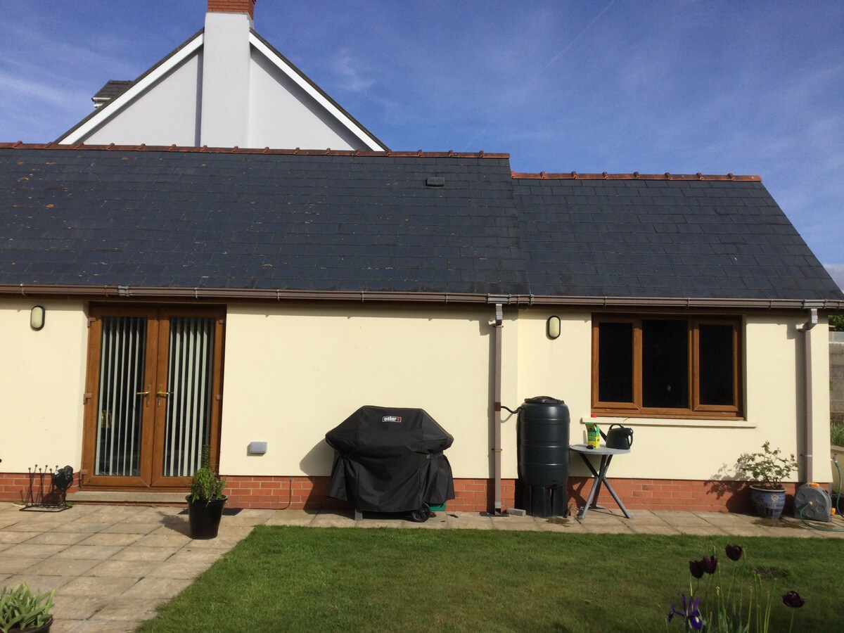 The exterior of the annex features a slate roof and light-colored walls, with large glass doors leading into the living space. A barbecue is positioned on the patio, along with a rainwater barrel and gardening tools. A well-maintained lawn surrounds the area.