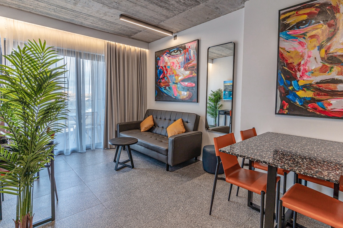 A spacious living area features a modern gray sofa with decorative pillows, complemented by a stylish dining table surrounded by four chairs. Vibrant artwork adorns the walls, and large windows allow natural light to fill the space while providing views of the outdoors.