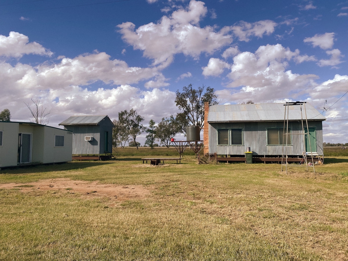 Two rustic buildings are situated in an open grassy area under a partly cloudy sky. A fire pit is visible in front of the larger building, and a clothesline stands nearby. Surrounding trees add to the rural landscape.