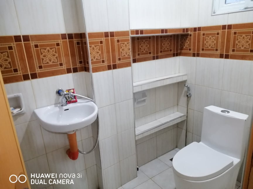 The bathroom features tiled walls in warm tones, complemented by a modern toilet and a wall-mounted sink. Shelving is provided for storage, enhancing functionality. Natural light enters through a small window, illuminating the clean, neutral color palette.