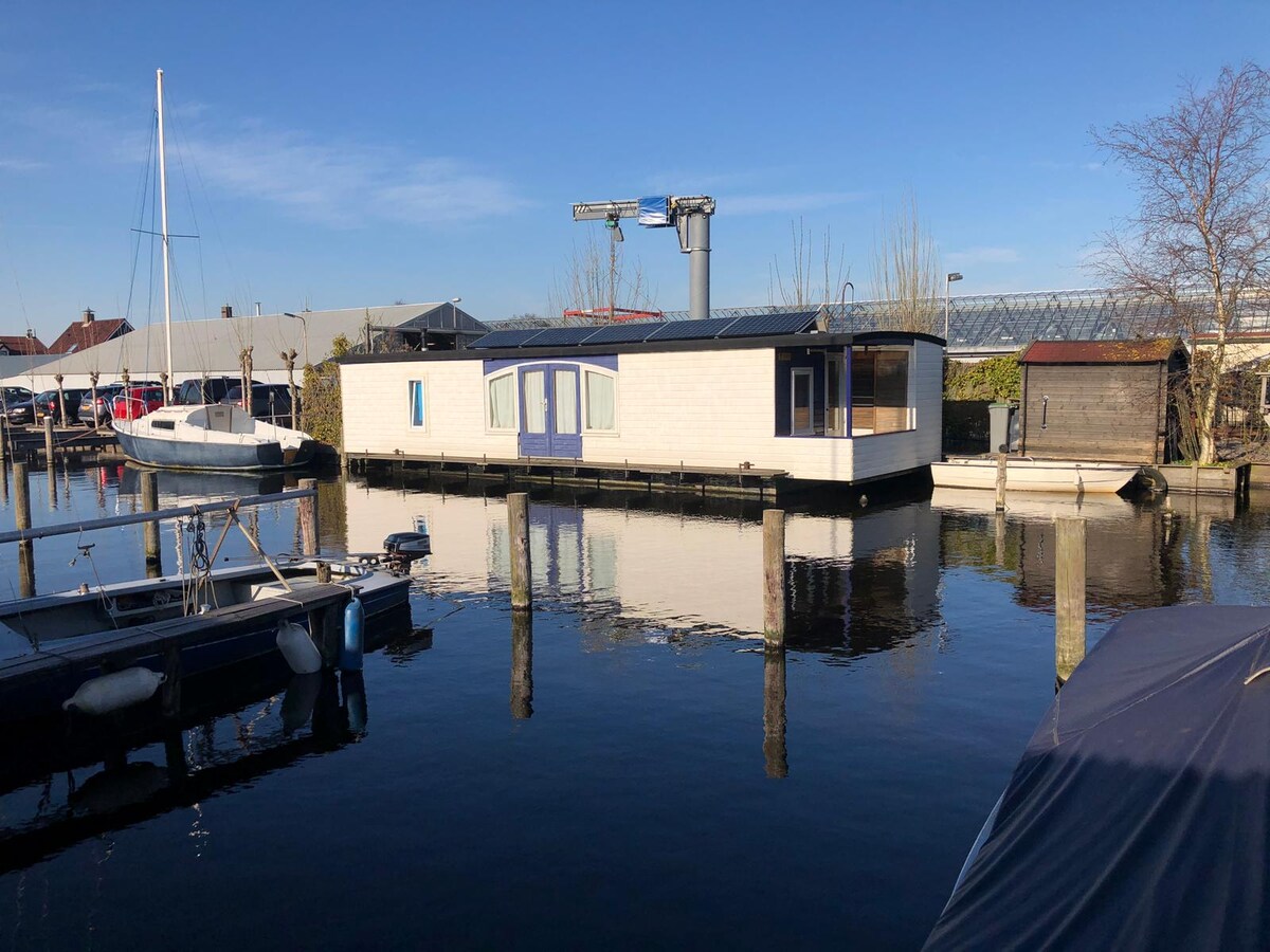 A modern waterhouse is depicted, featuring a sleek design and large windows. Its exterior is complemented by a clean, pale façade, surrounded by calm waters and gentle reflections. Nearby moored boats are visible, along with a hint of greenery in the background.