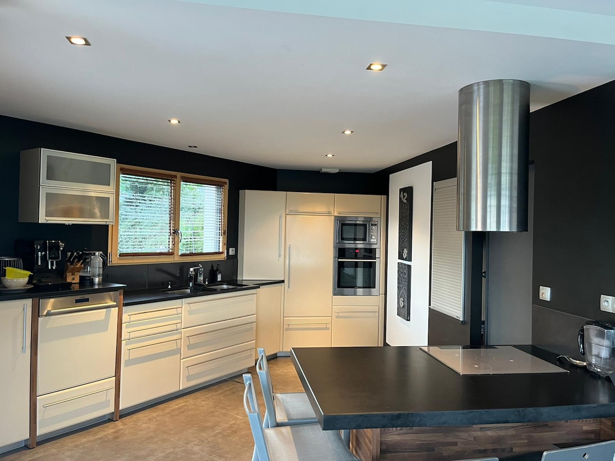 A modern kitchen features sleek cabinetry in neutral tones, complemented by a contrasting dark wall. A large island with a cooktop is highlighted, while natural light floods in through two large windows. Appliances include an oven, refrigerator, and dishwasher, creating a functional cooking space.