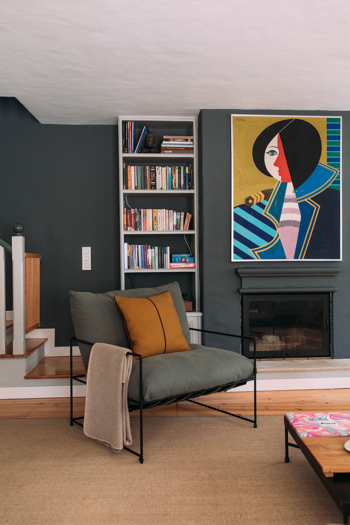A cozy corner of the living room features a chair with cushions and a throw draped over its side. A fireplace sits below a colorful artwork, while a bookshelf filled with books is positioned against the wall. Soft lighting enhances the inviting atmosphere.