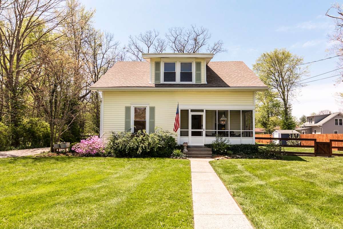 A charming farmhouse features a light yellow exterior with white trim and a welcoming front porch. Lush green grass surrounds the home, and colorful flowers can be seen near the entrance. A flag hangs by the porch, adding to the inviting presentation.
