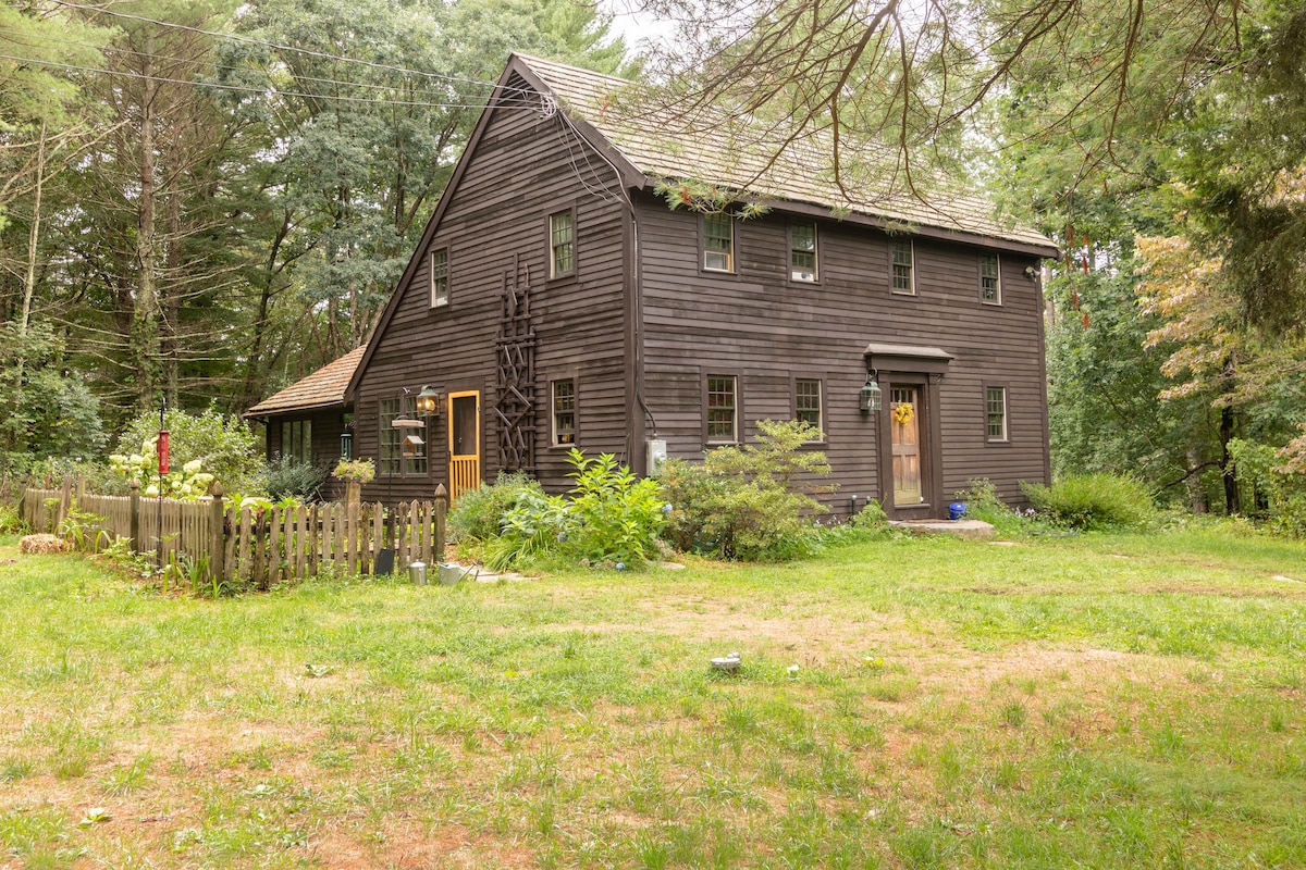 The exterior of a rustic wooden house is surrounded by lush greenery and trees. A small fenced area is visible near the entrance, where a welcoming front door can be seen. The building features windows and a gabled roof, set in a tranquil natural environment.