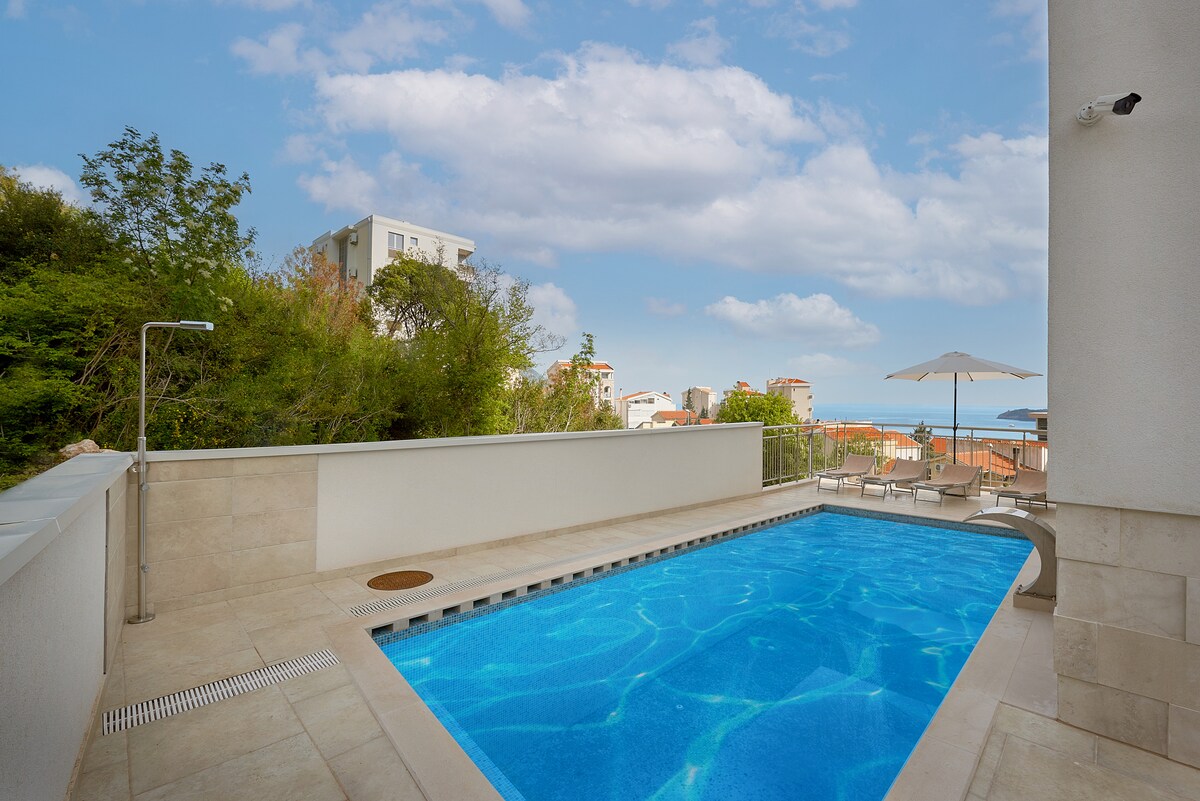 A private pool is framed by a modern tiled terrace, featuring clear blue water reflecting the sky. Sun loungers are positioned nearby under an umbrella, while greenery and distant buildings are visible in the background.