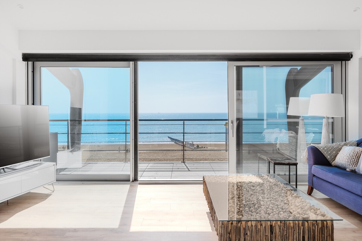 A spacious living area is illuminated by natural light, featuring a large smart TV and a glass coffee table with natural elements. Floor-to-ceiling windows offer unobstructed views of the beach and sea, with a balcony visible for outdoor enjoying.