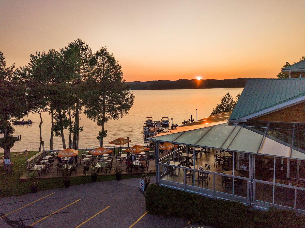 A lakeside dining area is viewed during sunset, with outdoor tables set up on a patio. The calm water reflects warm hues, while trees line the shore and boats are visible in the distance. A modern building with large windows frames the scene.