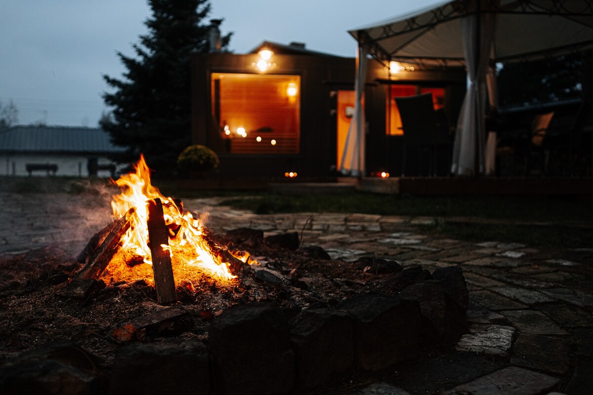 A fire pit is surrounded by a stone border, with flames gently rising from the center. In the background, a covered outdoor seating area with chairs and a table is partially visible, illuminated softly by overhead lights. Trees and an outdoor structure are seen beyond the fire.