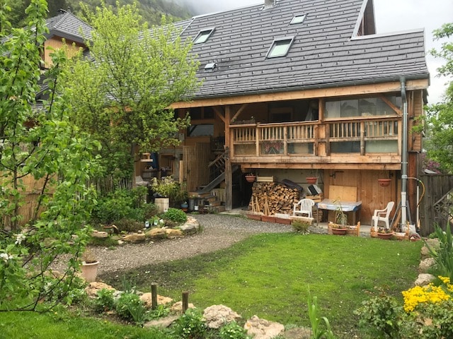 A charming wooden house is presented with a large covered terrace, adorned with a stack of firewood. A gravel path leads through a landscaped garden featuring greenery and flowers, inviting connection with nature. The house is set against a backdrop of gently sloping hills.