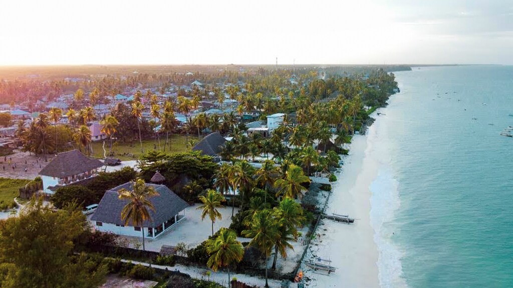 An aerial view captures the serene beachfront of the villa, surrounded by palm trees and gentle waves. The sandy shore complements the vibrant greenery of the nearby village, with traditional Makuti roofs visible. The tranquil ocean reflects soft hues of the setting sun.