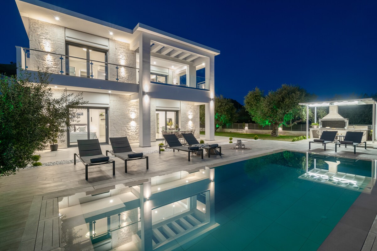 The villa is presented at night, featuring a well-lit exterior that highlights the modern design. A large pool reflects the surrounding lights, while sun loungers are arranged nearby. An outdoor dining area with a barbecue is visible, set against a backdrop of lush greenery.