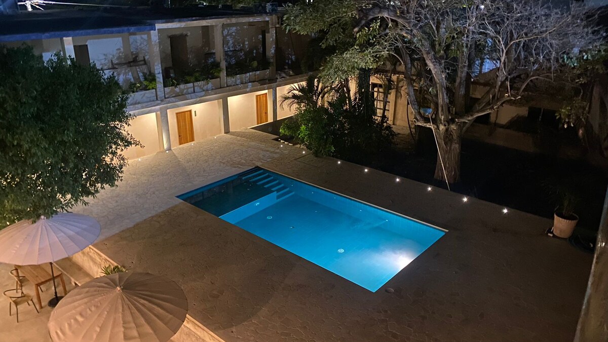 An inviting outdoor area features a spacious swimming pool surrounded by smooth stone tiles. Soft lighting accentuates the pool's clear water, while shaded seating areas with umbrellas offer spots for relaxation. Lush greenery and trees provide a natural backdrop to the serene space.
