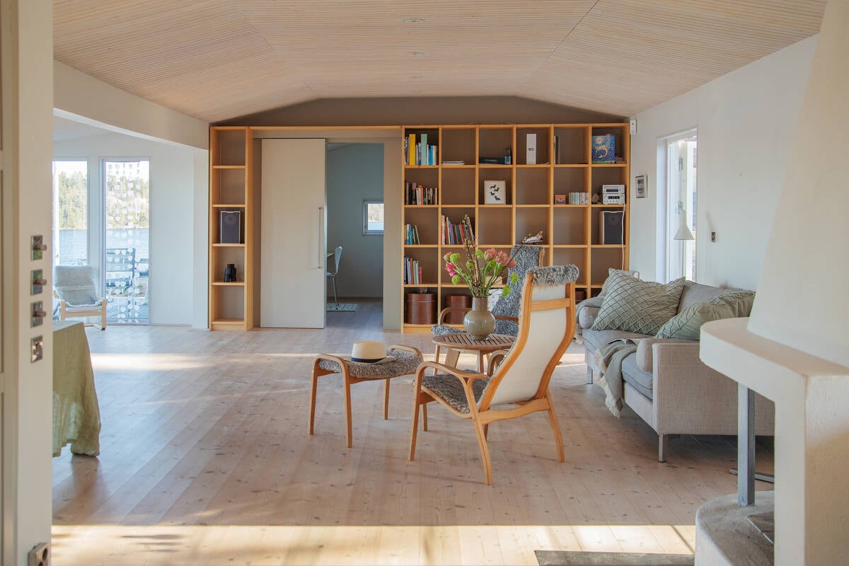 A spacious living area is characterized by natural wood flooring and soft light from large windows. A comfortable sofa, an armchair, and a coffee table create an inviting seating arrangement. Bookshelves provide functional storage, while decorative elements add subtle interest to the space.