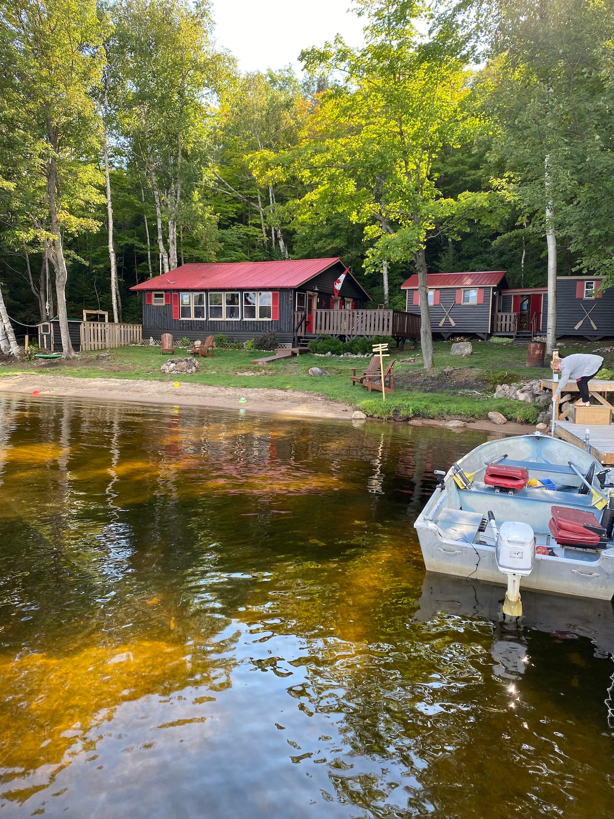 A cozy cottage with a red roof is nestled among trees, situated near a calm water area. A small sandy beach area is visible, and a boat is anchored nearby. The surrounding landscape features greenery and a peaceful setting, ideal for relaxation.