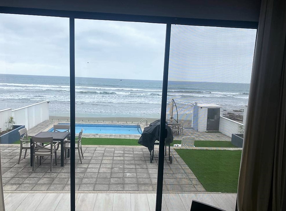 A panoramic view of the ocean can be seen through large sliding glass doors. The outdoor space features a patio area with a dining table, a pool, and an expanse of green grass. Waves gently lap against the shore, enhancing the tranquil environment.