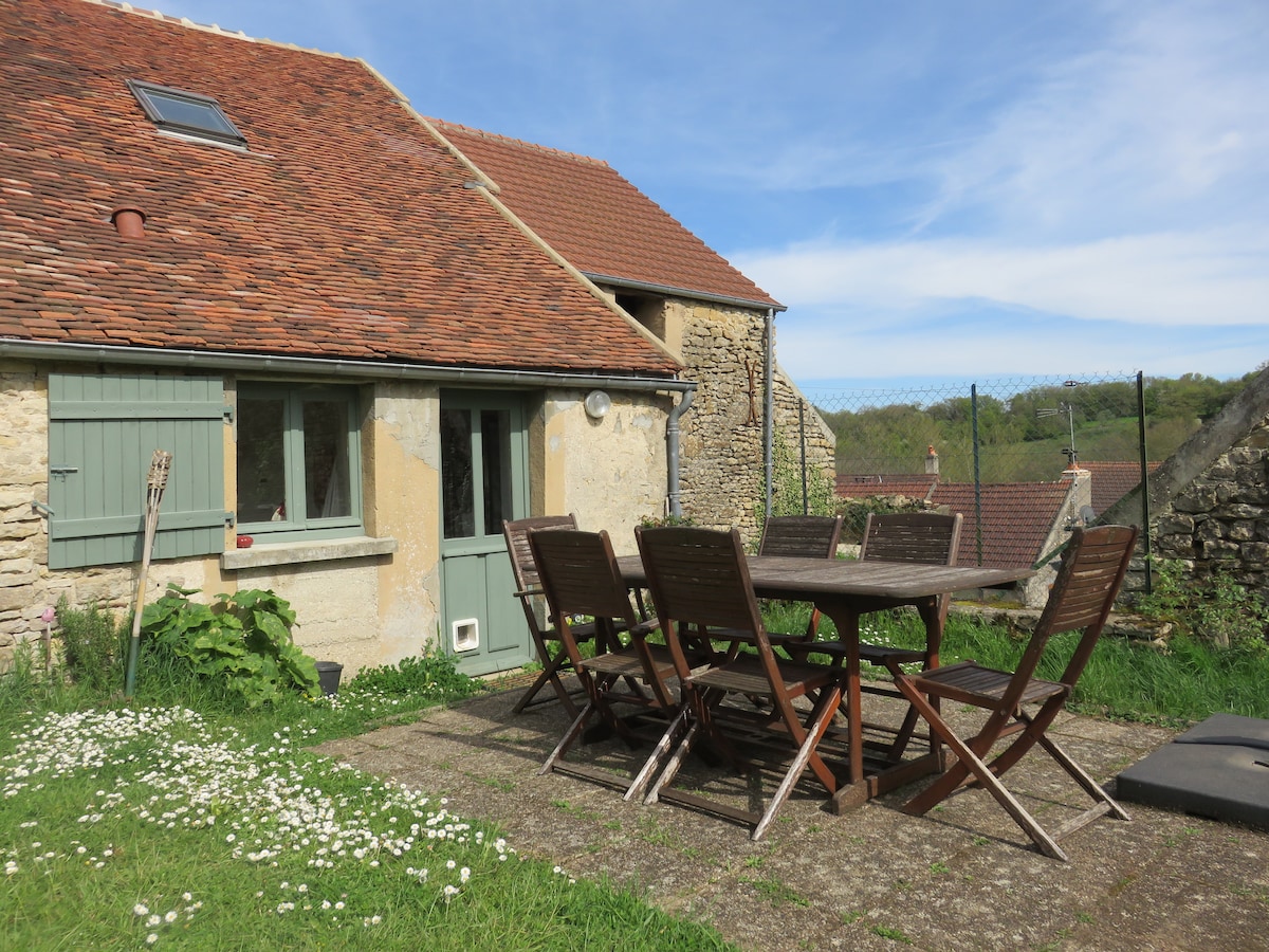 An outdoor terrace area includes a large wooden table surrounded by several wooden chairs, set on a grassy patch. The rustic stone building features green shutters and a slanted roof, with trees visible in the background under a clear blue sky.
