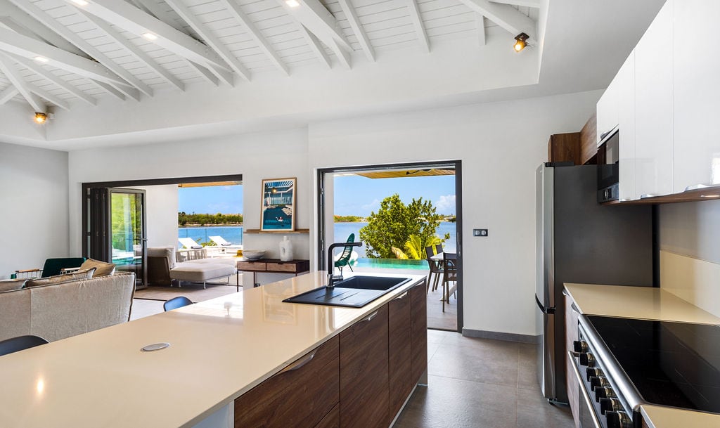 A modern kitchen is highlighted with sleek cabinetry and a central island. Large sliding doors open to a scenic patio overlooking the lagoon. Natural light fills the space, showcasing a well-equipped kitchen area with stylish appliances and a spacious layout.