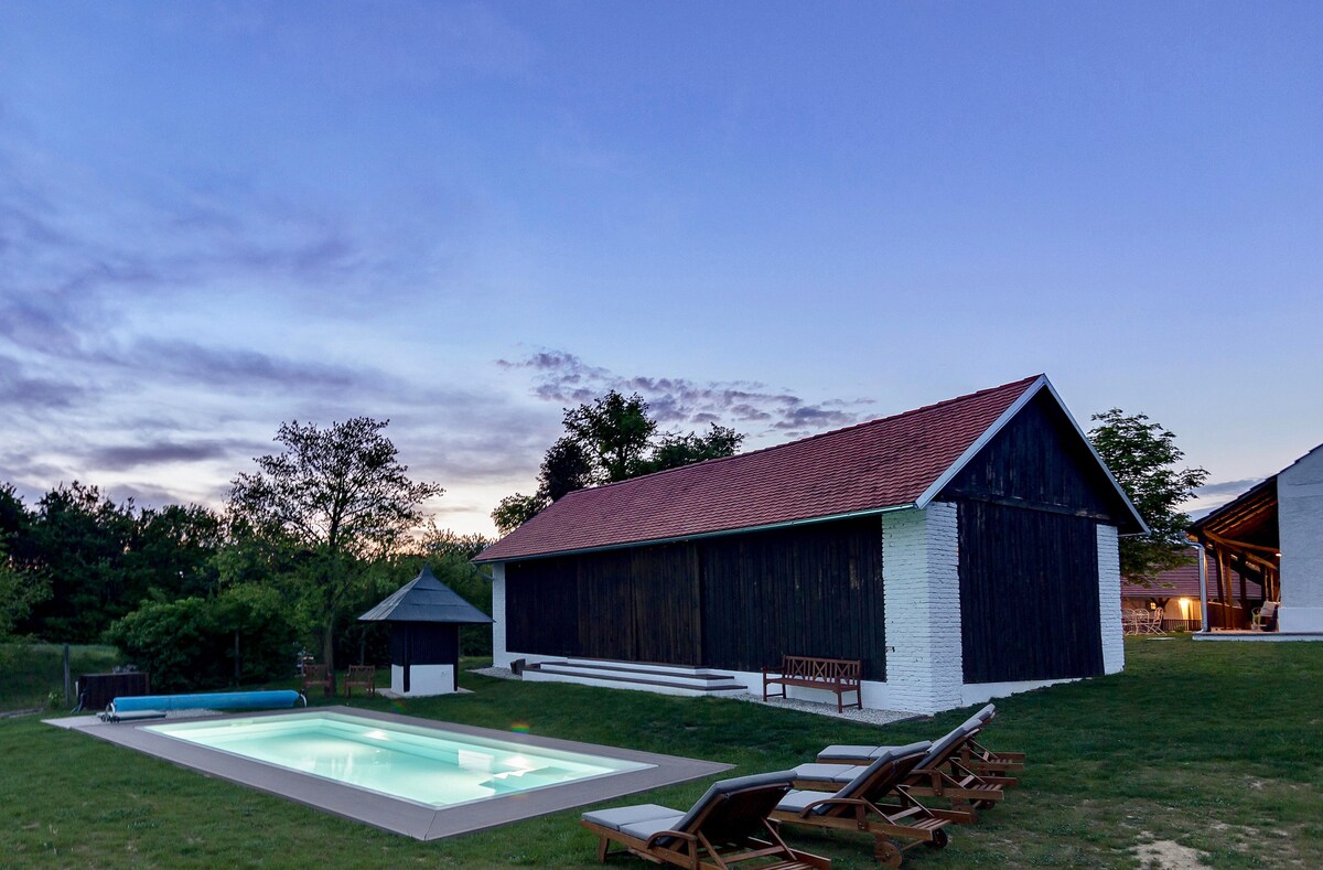 The image features a serene outdoor pool area beside a rustic building with a wooden facade. Lounge chairs are arranged nearby on a grassy lawn, surrounded by trees. The sky showcases soft twilight hues, complementing the tranquil setting.