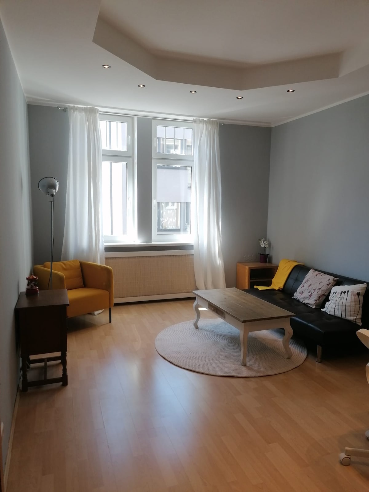A well-lit living area is featured, with large windows allowing natural light to enter. A black sofa and a yellow armchair provide comfortable seating. A round area rug lies on the light wooden floor, complemented by a small coffee table and side table with decorative items.