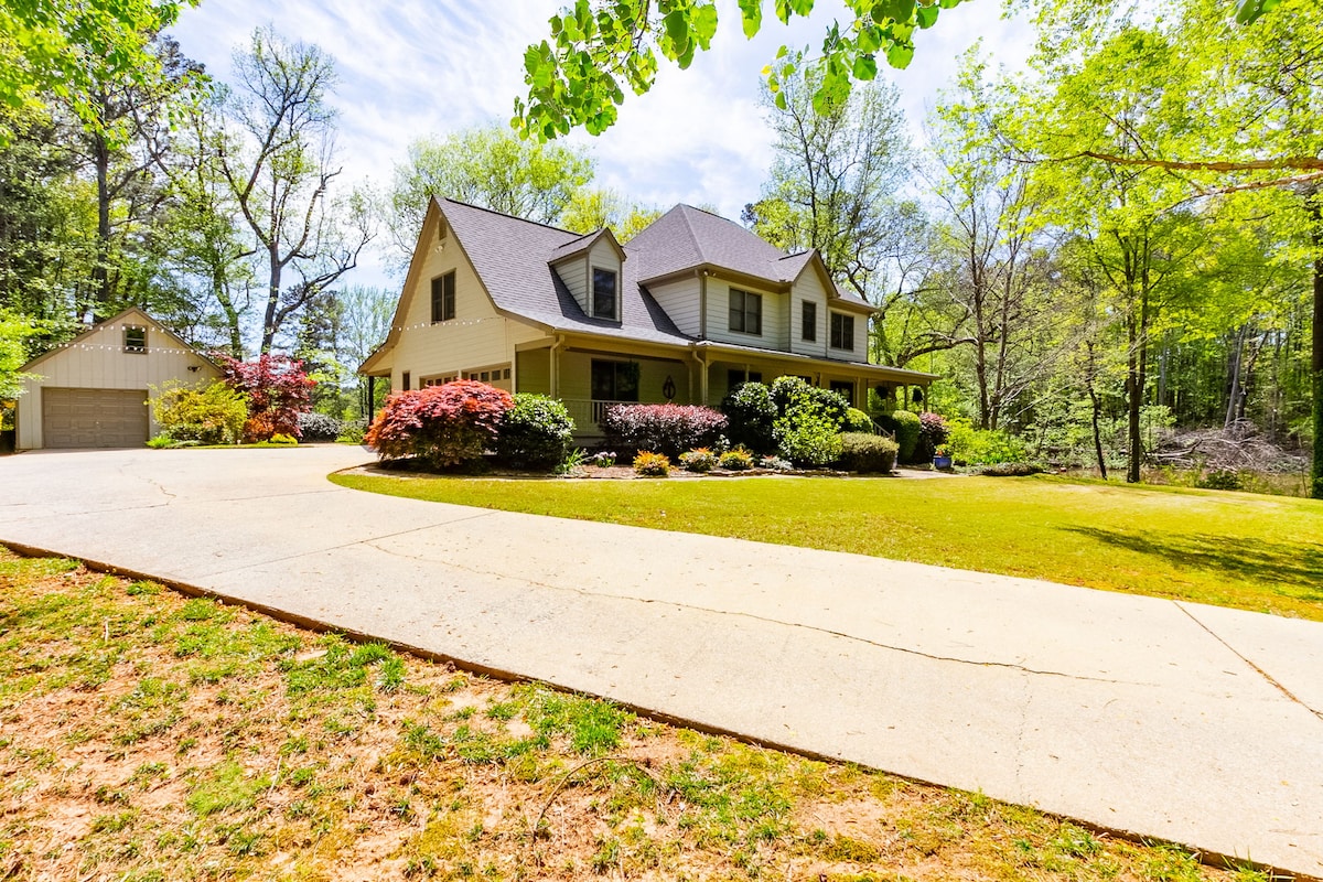 A large home is set within a lush landscape, showcasing well-maintained grass and flowering bushes. A spacious driveway leads up to the inviting entrance, and a separate garage is visible to the left, surrounded by trees and greenery.