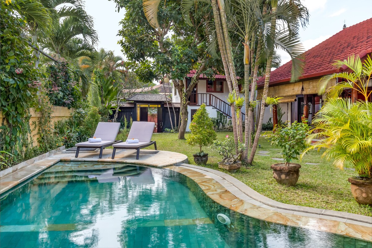 A tranquil infinity pool is surrounded by lush gardens, with two lounge chairs positioned nearby. Tropical plants and palm trees create a serene environment, complemented by the warm tones of the villa's exterior visible in the background.