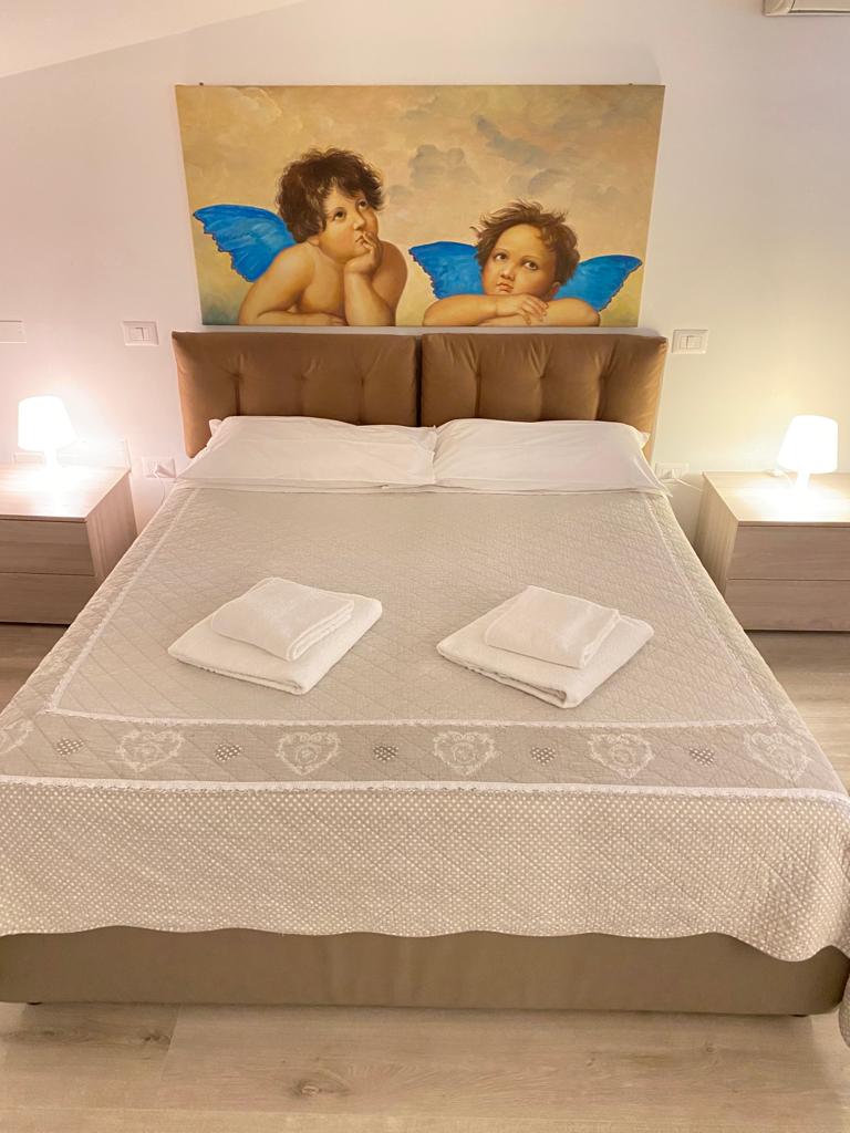 A spacious bed is centrally placed, topped with a light-colored blanket. Two neatly folded towels are positioned at the foot of the bed. On either side, matching bedside tables with lamps provide balanced lighting. The wall behind features a mural of two cherubs with blue wings.