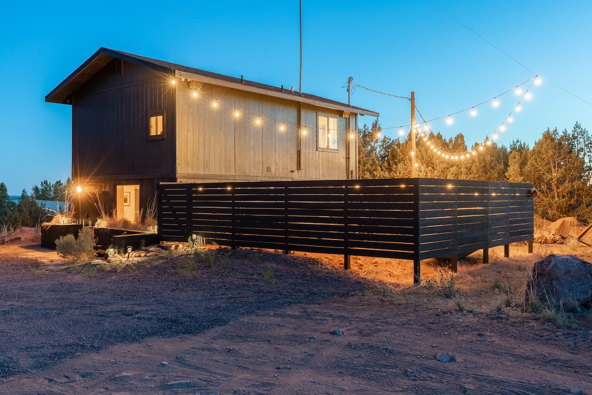 The exterior of the Radio Tower is illuminated by string lights, creating a welcoming ambiance. The wooden structure is surrounded by natural desert terrain, with a patio area visible that includes a private hot tub. The stars begin to emerge in the evening sky.