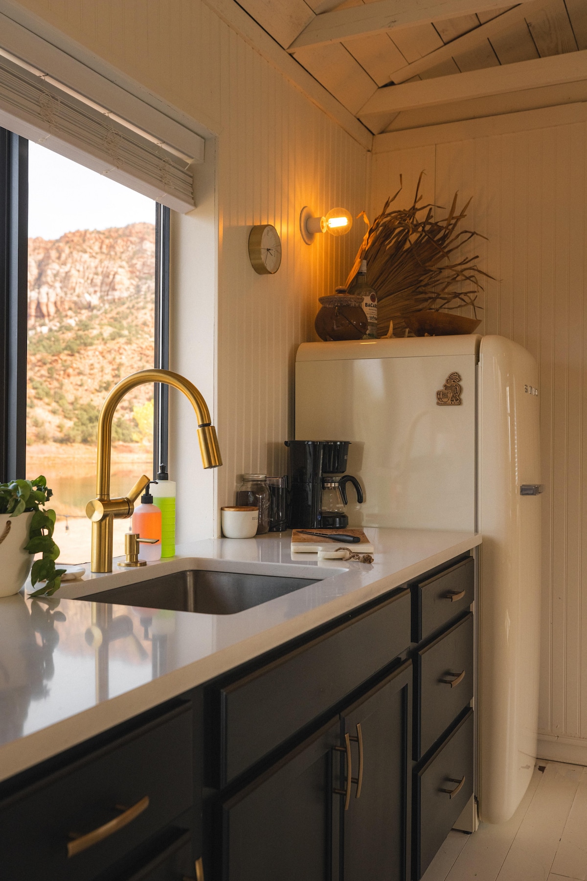 The kitchen features a striking view of the surrounding mountains through a large window, with a mix of modern and vintage elements. A gold faucet stands above a stainless steel sink, while a white retro refrigerator complements the dark cabinetry. Coffee maker and utensils are organized on the countertop.