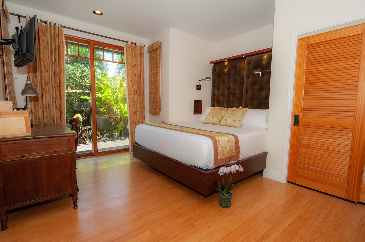 A spacious studio suite features a California King bed with elegant high thread count linens. Natural light filters through large windows, revealing tropical greenery outside. A wooden cabinet and bedside lamp add functionality, while a textured headboard complements the room's calming aesthetic.