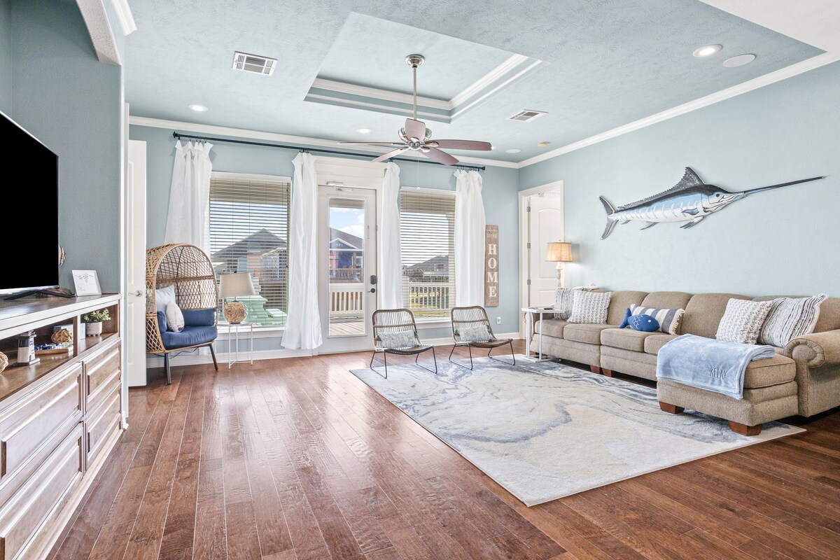 A spacious living area features wooden flooring and a large area rug. A comfortable sofa is complemented by two accent chairs. Natural light streams through large windows, and a decorative fish hangs on the wall. The room invites relaxation with its airy feel.