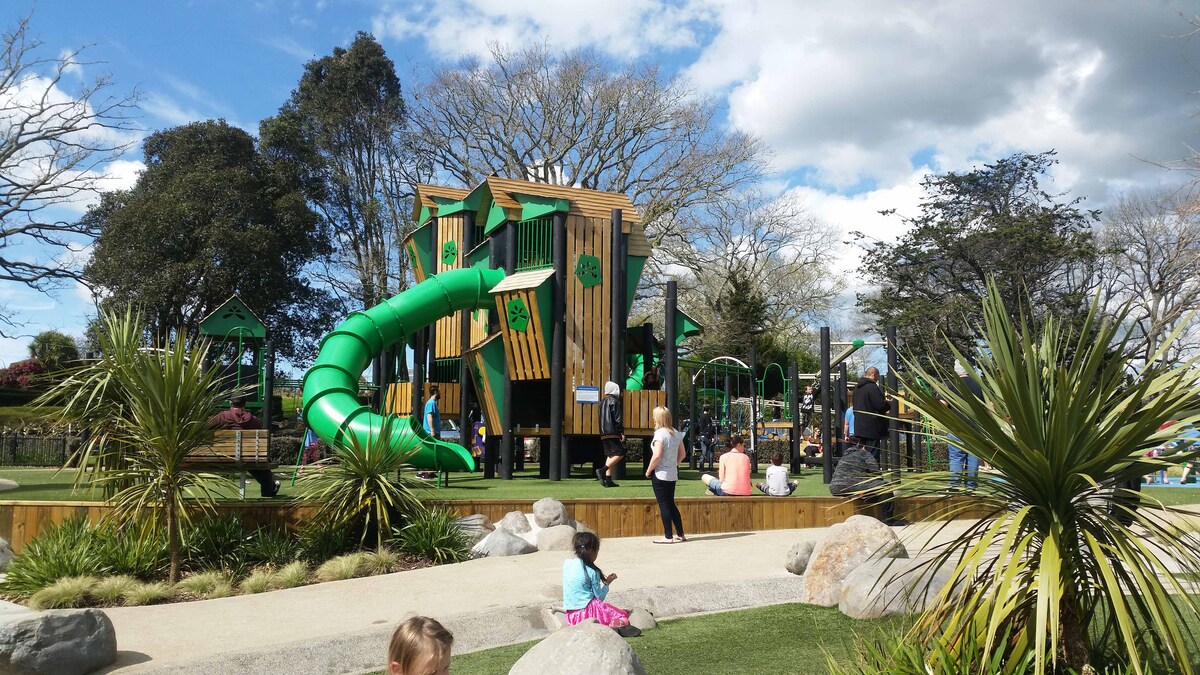 A vibrant playground features a large wooden structure with multiple levels, incorporating a green slide and climbing elements. Children are seen playing in various areas, with landscaped grass and boulders surrounding the space, while shaded trees provide a natural backdrop.