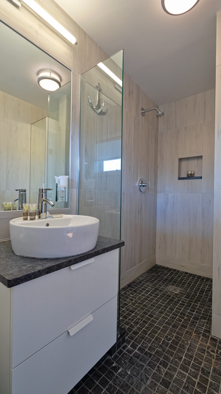 A modern bathroom is displayed, featuring a sleek vessel sink atop a minimalist white cabinet. The shower area includes glass walls and a handheld showerhead, with light-colored wood panels complementing the dark stone flooring. Natural light is welcomed through a small window.
