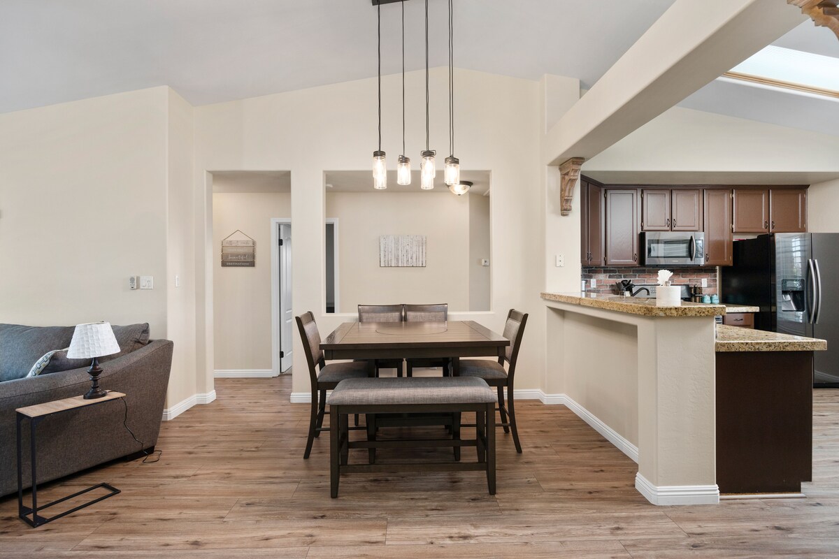 A modern dining area features a rectangular table surrounded by six chairs, complemented by a light fixture overhead. A spacious living area is visible in the background, with a comfortable gray couch and kitchen accessible nearby, showcasing dark cabinetry and stainless steel appliances.
