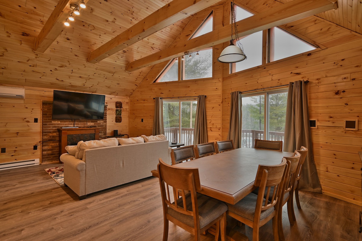 A spacious living area is highlighted by natural wood paneling, featuring large windows that invite ample light. A comfortable sofa faces a mounted television, while a dining table with multiple chairs offers seating for group meals. The high ceilings create an open atmosphere.