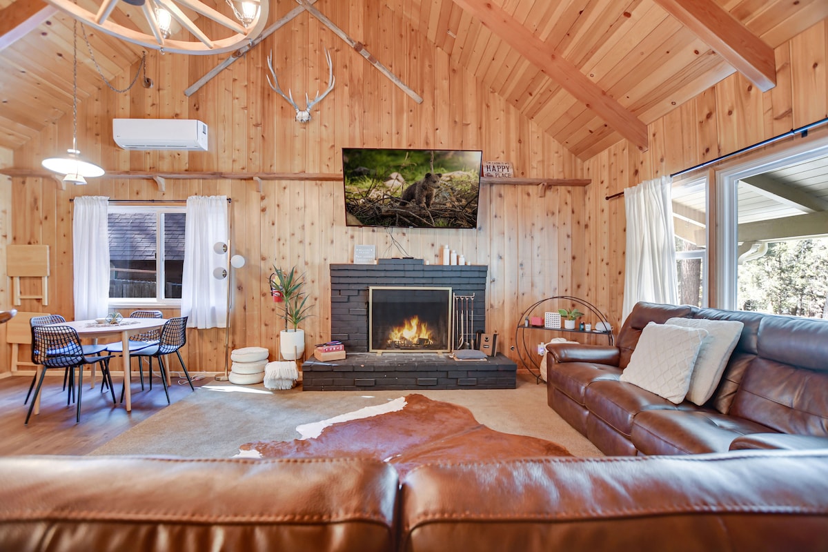 A warm and inviting living area features a wood-burning fireplace with a flat-screen TV mounted above. The vaulted ceiling enhances the rustic charm, while a dining table and chairs are positioned nearby. Large windows offer views of the outdoors, with light streaming into the space.