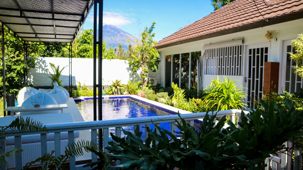 The image showcases a serene outdoor area featuring a pool surrounded by lush greenery. Mount Agung is visible in the background. A shaded seating area is present, providing an inviting space to relax. The garden's vibrant foliage enhances the tranquil atmosphere.