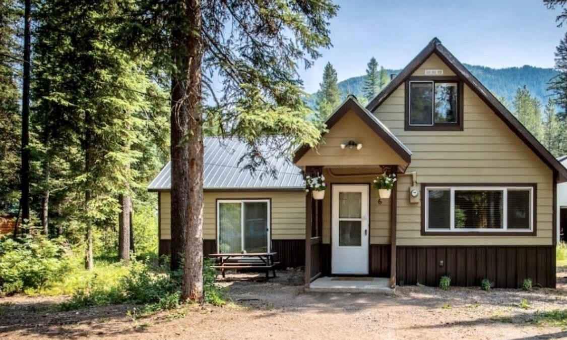 The cabin's exterior features a peaked roof and a welcoming front porch. Surrounded by tall pine trees, a picnic table is positioned near the entrance. Large windows reflect the natural surroundings, inviting daylight into the space.