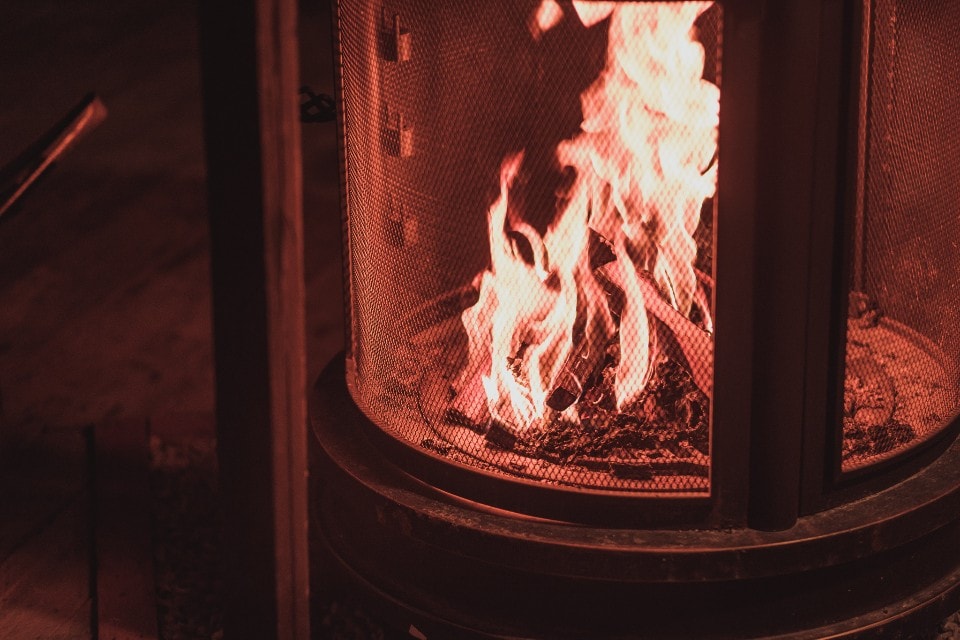 A warm fire burns inside a metal fireplace, surrounded by a mesh screen. The flames flicker in shades of orange and red, casting a soft glow on the surrounding area. The ambiance suggests a cozy atmosphere, perfect for relaxation.