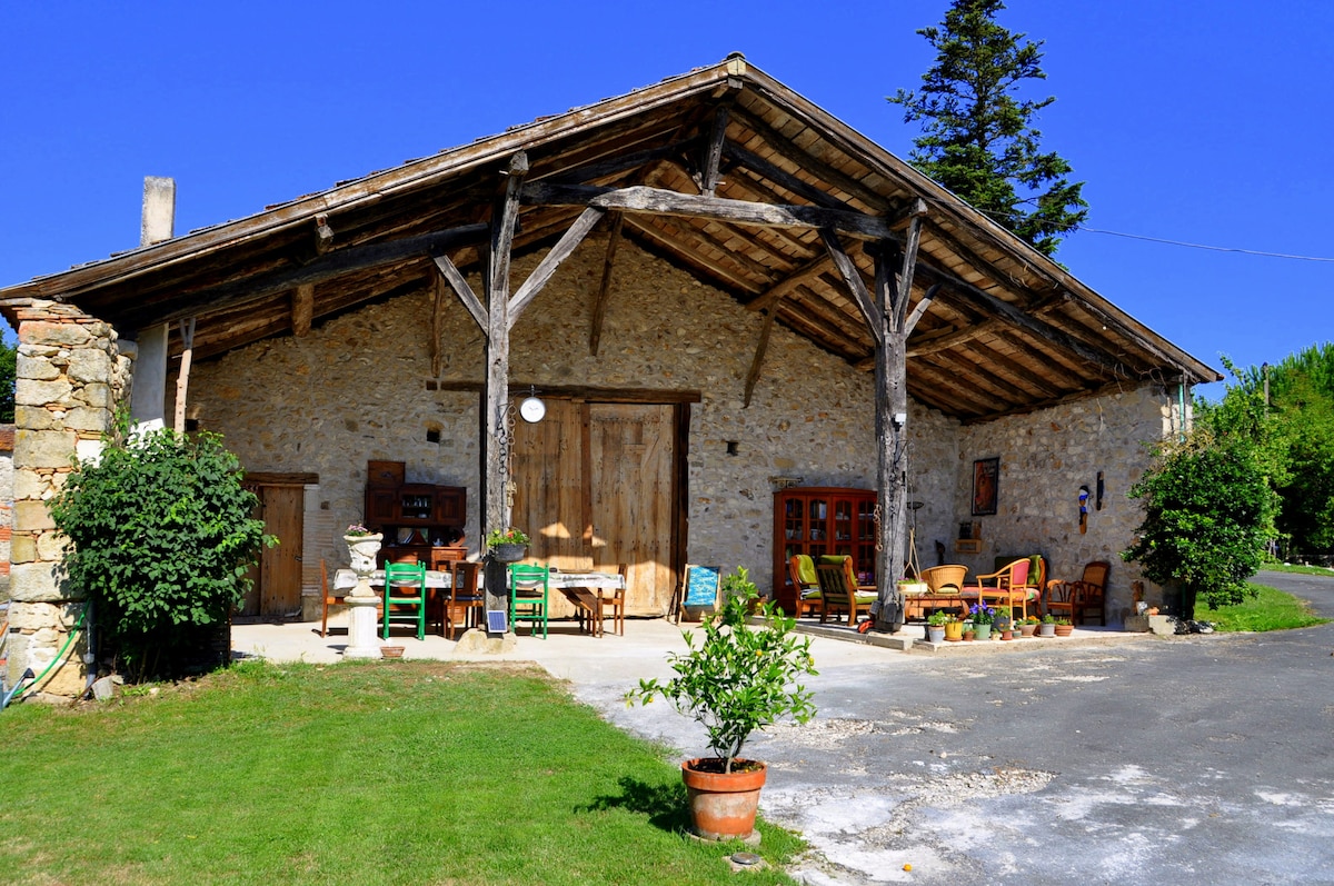 The image displays a rustic barn featuring a large wooden awning supported by sturdy beams. Outdoor seating areas with colorful chairs and tables are arranged under the awning, surrounded by greenery. A stone façade complements the warm, inviting atmosphere of the space.