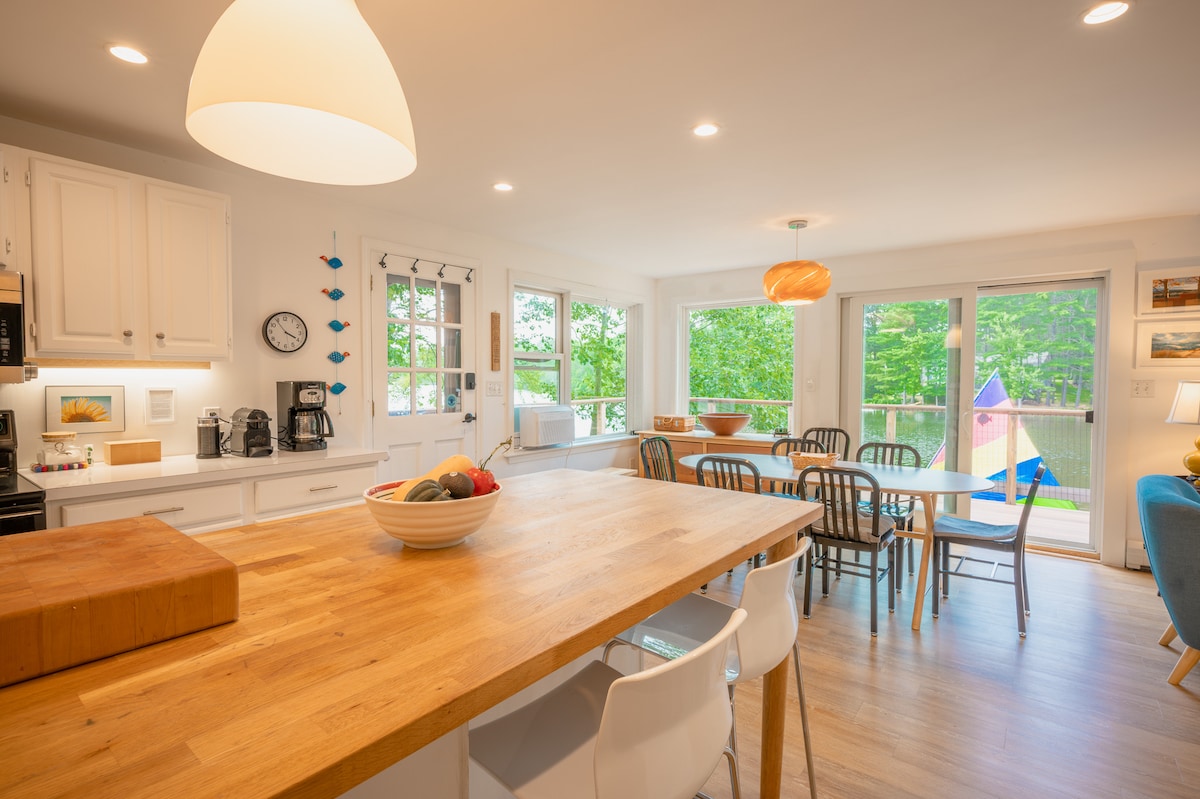 A bright and open kitchen-dining area features a large wooden table with an inviting bowl of fruit at its center. Multiple chairs surround the table, and large windows provide natural light and views of the outdoors, with colorful outdoor decor visible.