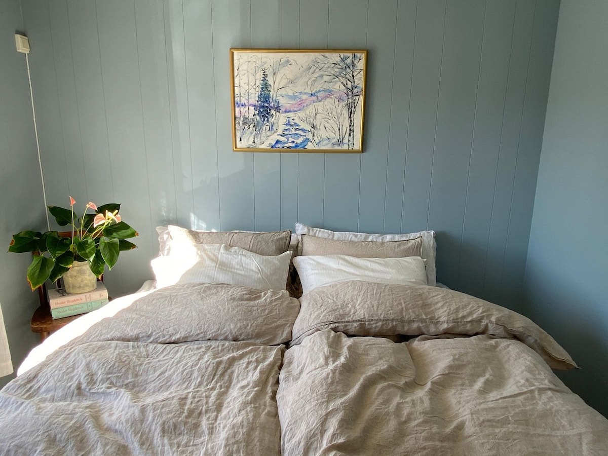 A spacious bed is dressed with soft beige linens, complemented by white pillows. A decorative painting depicting a serene landscape hangs above the bed. Natural light illuminates the room, highlighting a small green plant on a bedside table.