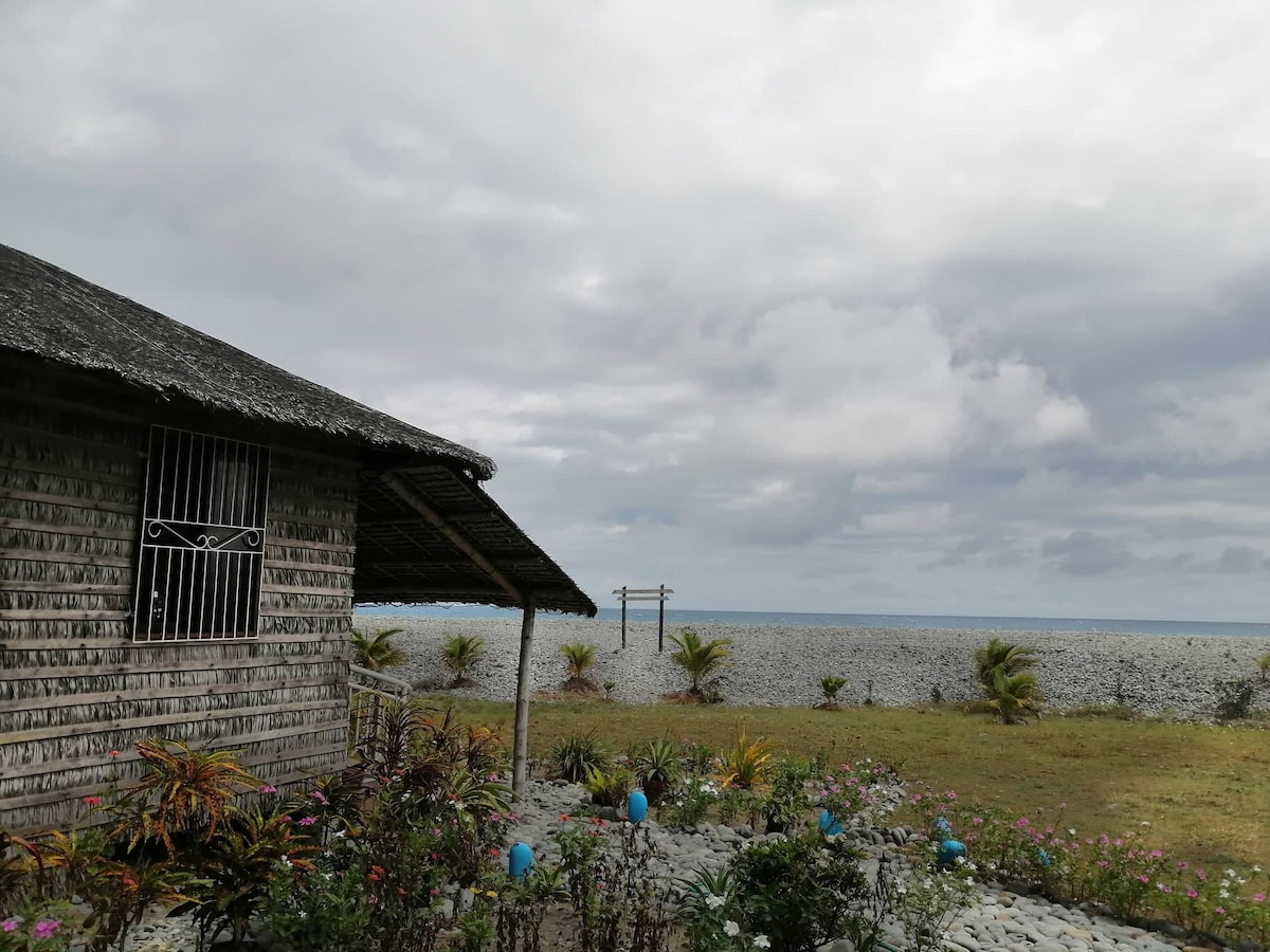A cottage constructed with natural materials is shown beside a landscaped garden, featuring colorful plants and decorative stones. The view extends to a pebble beach and the ocean under a cloudy sky, enhancing the sense of tranquility in the setting.