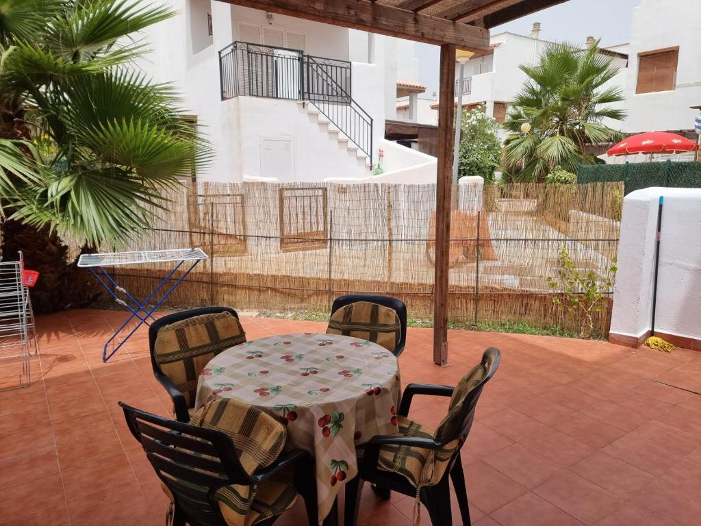 A shaded outdoor patio area features a round table set with a floral tablecloth and four black chairs. Lush palm trees are visible, alongside a clothes drying rack. The background shows a residential building with a staircase and a fenced garden area.