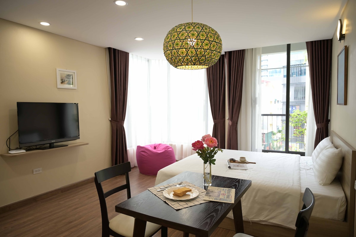 A welcoming living area features large windows allowing natural light to fill the space. A cozy bed is accompanied by a small dining table set for two. A vibrant pendant light adds a touch of color, while a television is mounted on the wall.