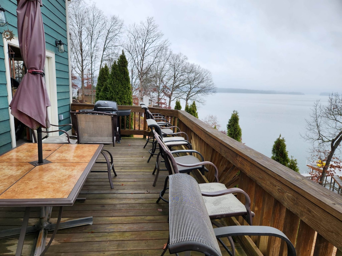 A spacious wooden deck features multiple chairs and a table, with a grill positioned towards the edge. The surrounding trees offer a glimpse of the serene lake through the overcast sky, emphasizing the peaceful atmosphere of the space.