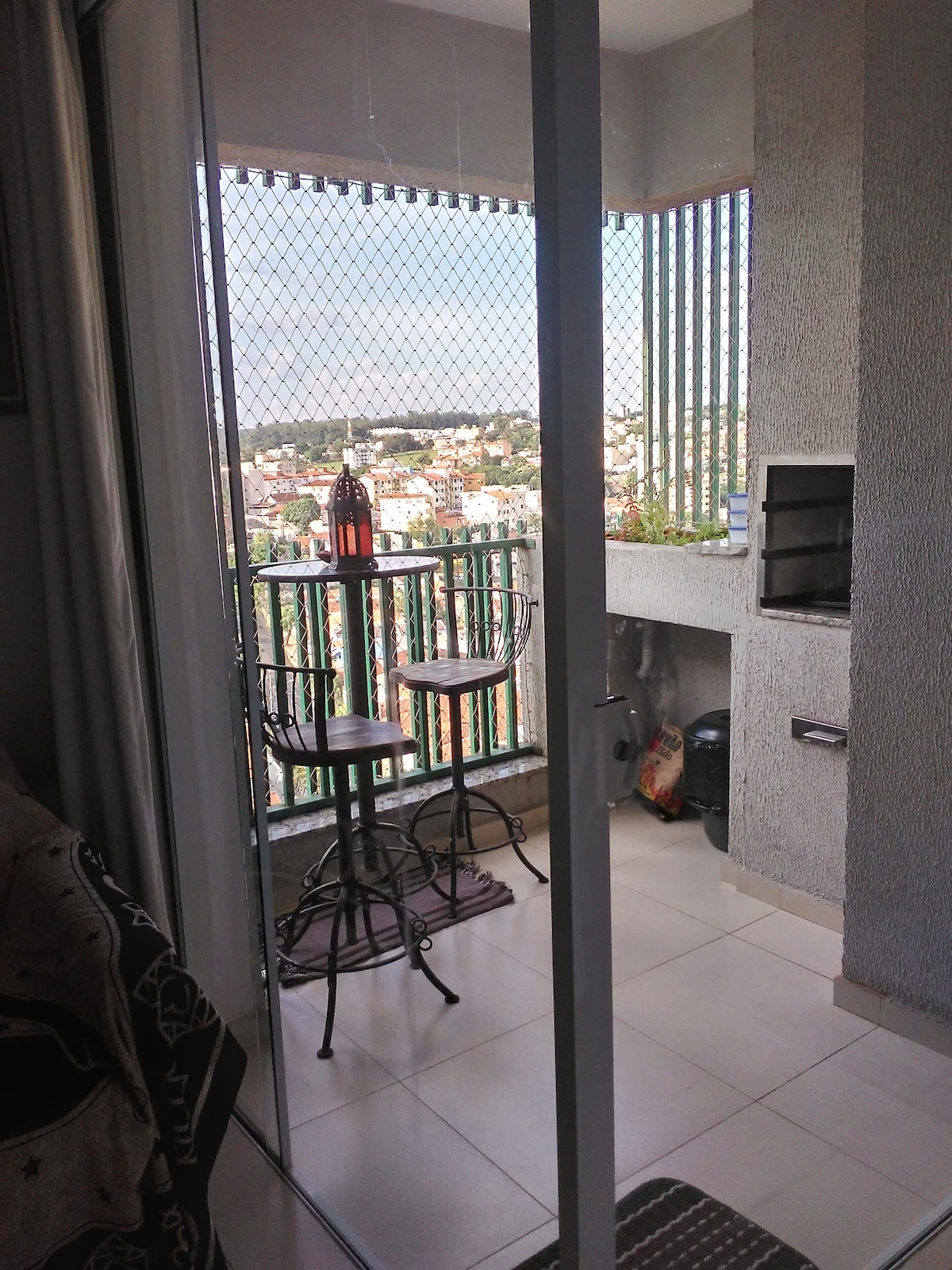 The balcony area features a small bar table with two high stools, providing a space for casual dining or relaxation. A grill is positioned adjacent to the table. The view includes urban scenery, framed by a metal railing, allowing natural light to fill the space.
