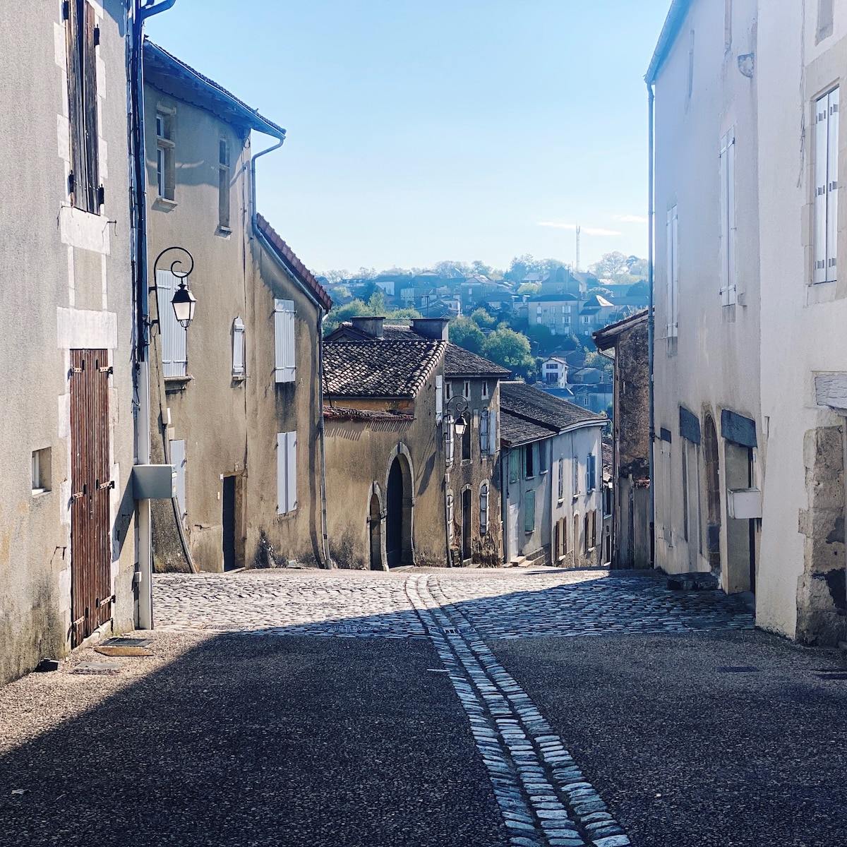 A narrow, cobbled street is framed by historic buildings featuring various shades of beige and white. The gentle slope of the street leads to distant hills, providing a backdrop of greenery under a clear blue sky. A streetlamp is visible, adding a charming touch to the scene.