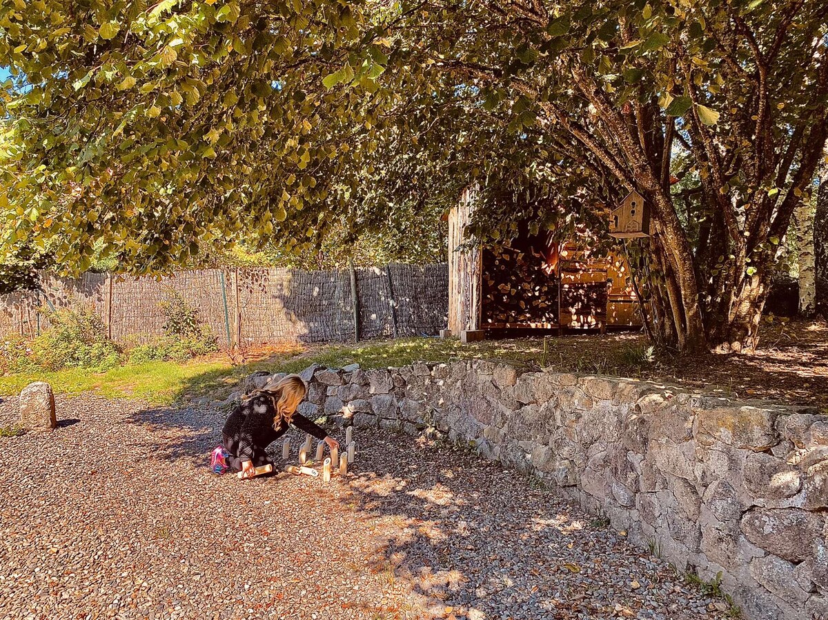 A child is seen playing on a gravel path beneath a large tree, surrounded by dappled sunlight. A rustic building is partially visible in the background, and a stone wall borders the area, creating a calm and inviting outdoor space.