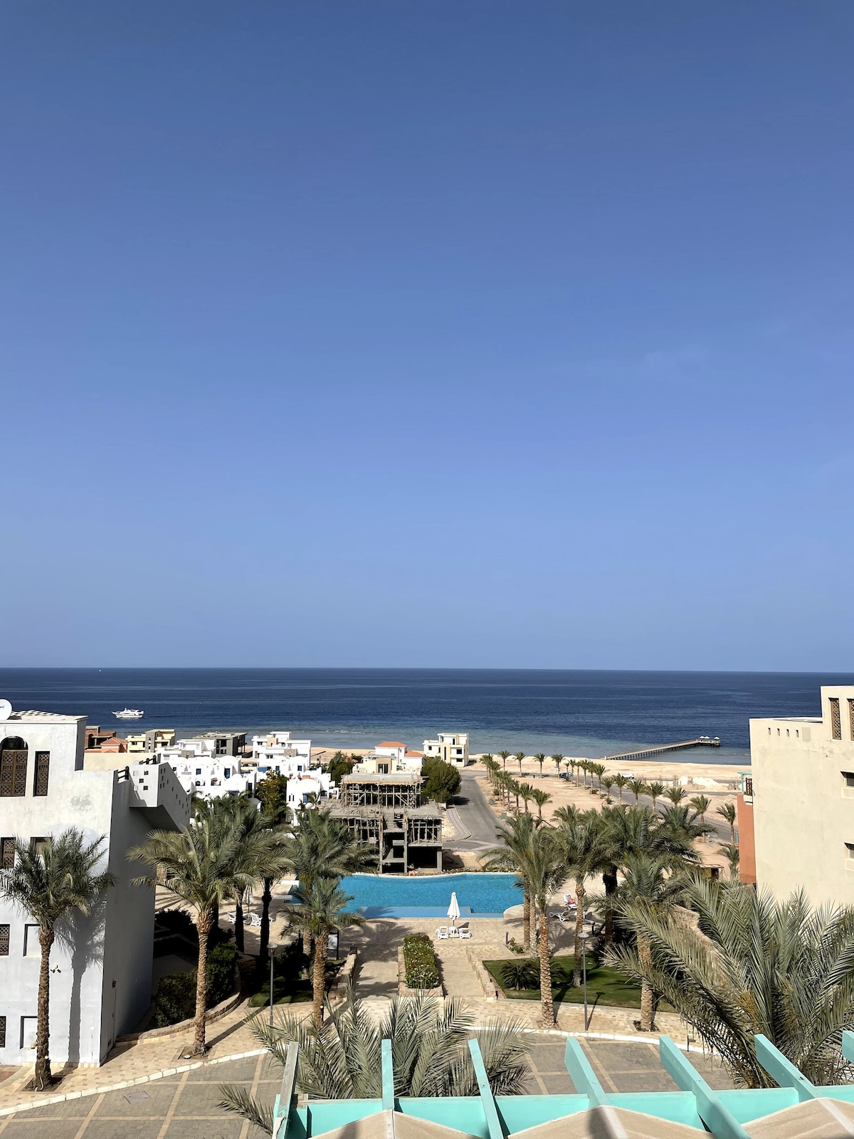 An expansive view overlooks a tranquil sea and a landscaped area featuring palm trees and a swimming pool. The clear blue sky complements the serene water, creating a harmonious coastal scene. Nearby buildings are positioned to create an inviting atmosphere.
